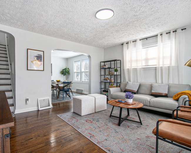The image shows a cozy living room with a gray sofa, two ottomans, a wooden coffee table, and a staircase visible in the background.