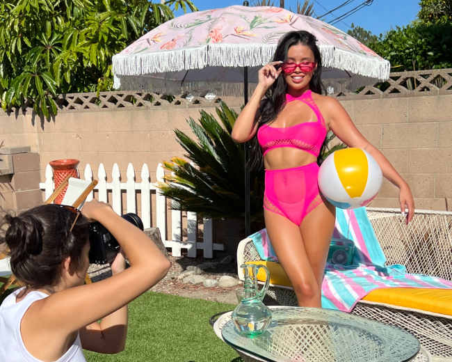 A photographer captures a model posing in a pink swimsuit by a poolside with a beach ball while a colorful umbrella provides shade.