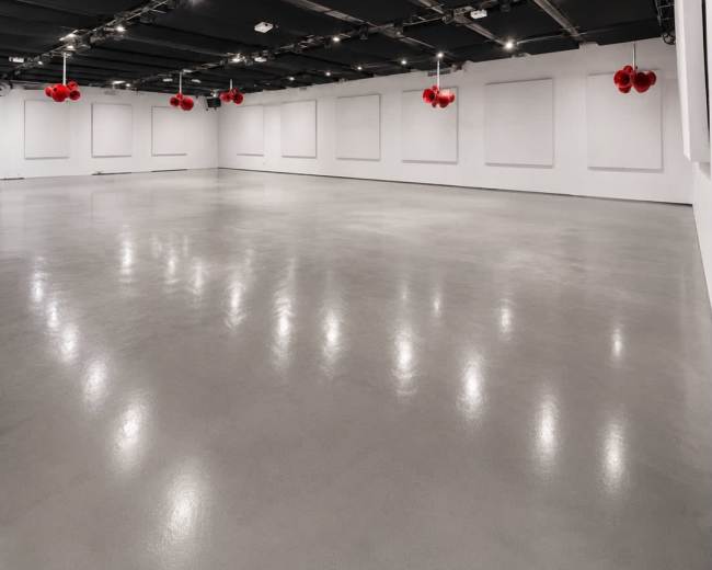 The image shows a stark, empty gallery space with polished concrete flooring and large white walls featuring several suspended red orbs.