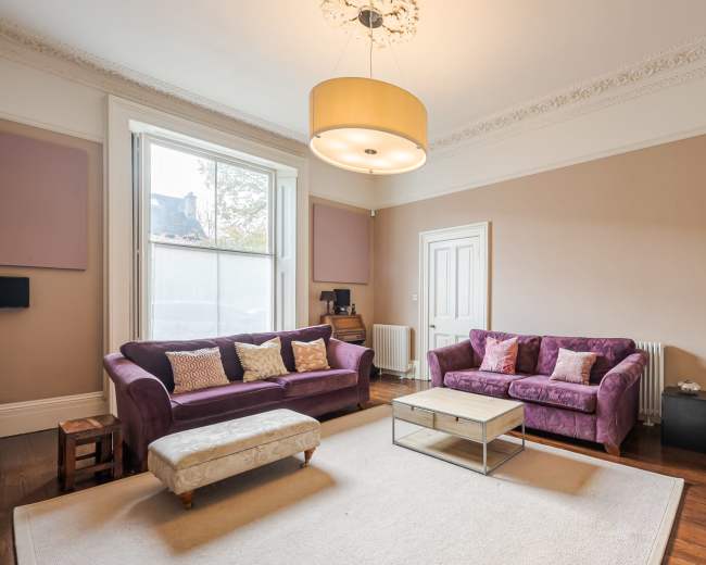 The image shows a spacious living room with purple sofas, a light fixture overhead, a wooden floor, and a coffee table in the center.