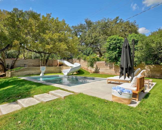 The image shows a backyard with a swimming pool, a water slide, lounge chairs, and a basketball hoop, surrounded by greenery.
