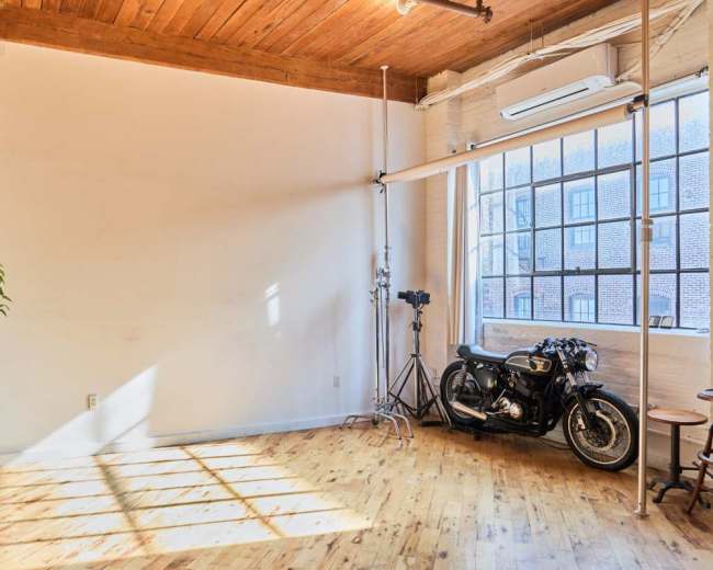 The image shows a spacious, brightly lit room with wooden flooring, large windows, and a vintage motorcycle parked against a wall.