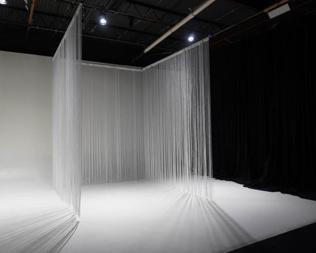 The image shows a minimalist studio space with sheer, vertical curtains dividing the area against a backdrop of white and black walls.