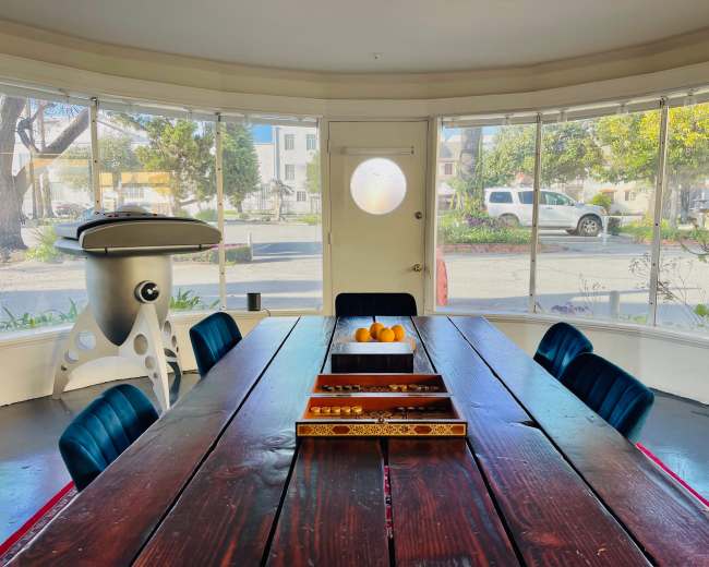 A large wooden dining table with a decorative tray of fruits is set in a bright room surrounded by large windows.