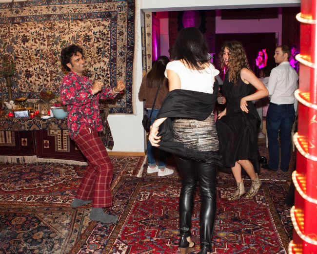 A group of people dance at a festive gathering surrounded by colorful rugs and intricate wall hangings.