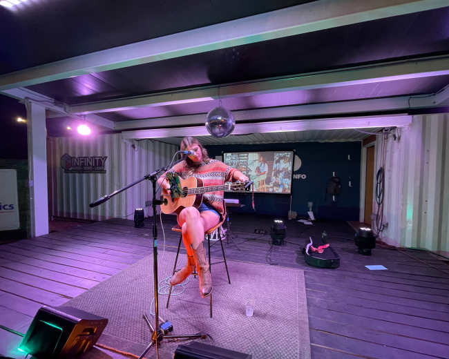 A woman plays an acoustic guitar while seated on a stool in a decorated outdoor stage area, with a video screen displaying images behind her.