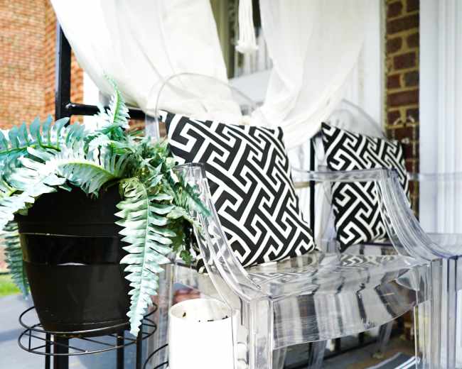 A transparent acrylic bench with patterned black and white cushions sits alongside a potted plant on a covered porch.