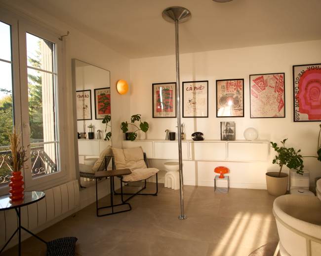 The image depicts a bright room featuring a pole, modern furniture, and framed artwork on the walls, complemented by indoor plants.