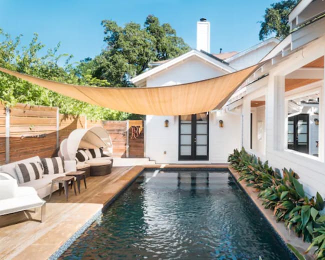 The image shows a modern outdoor living space featuring a rectangular swimming pool surrounded by plants, a cozy seating area, and a large shade sail overhead.