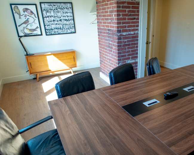 A conference room features a large wooden table with black chairs, a brick pillar, and a wooden sideboard against the wall adorned with artwork.