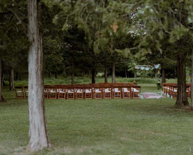 A set of wooden chairs is arranged in rows for a ceremony in a grassy area surrounded by trees.