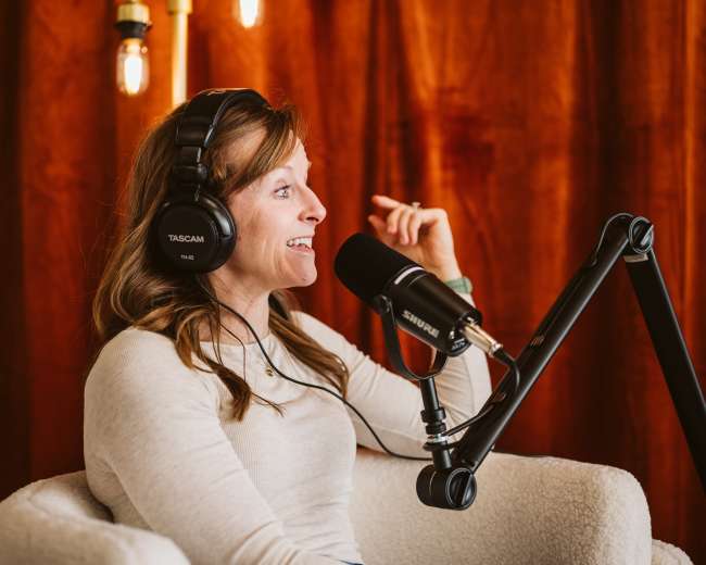 A woman with headphones smiles while speaking into a microphone in a cozy recording studio setting.