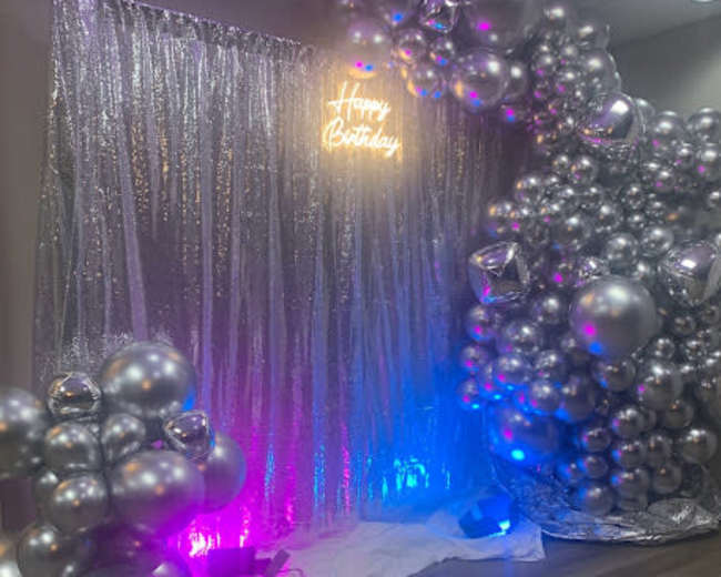 A decorative backdrop for a birthday celebration, featuring a silver curtain, neon "Happy Birthday" sign, and an arrangement of metallic balloons.