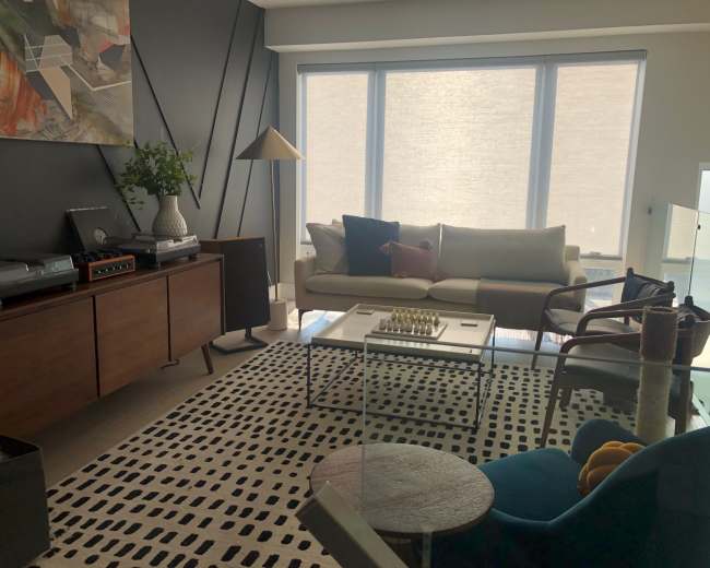 The image depicts a modern living room with a mix of furniture, including a sofa, a coffee table with a chess set, and a patterned rug, all set against a backdrop of large windows and a dark accent wall.