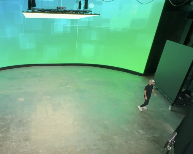 A person stands on a large, empty stage with a curved green screen backdrop, while another person works at a desk with equipment on the side.