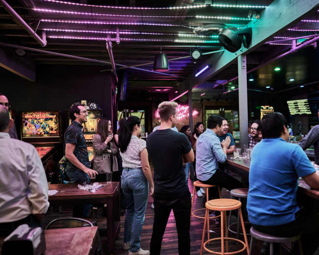 A group of people socializes at a bar with neon lights and arcade games in the background.