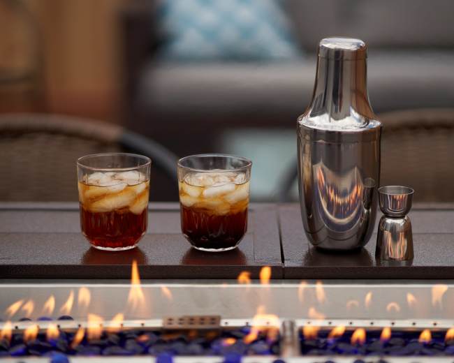 Two glasses filled with ice and a dark liquid sit beside a metal shaker and jigger on a table with flames and blue stones in the foreground.