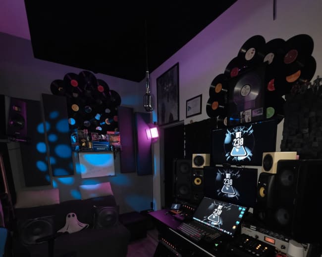 The image shows a music studio featuring a desk with computer monitors, various speakers, and a cozy seating area adorned with vinyl records on the walls.