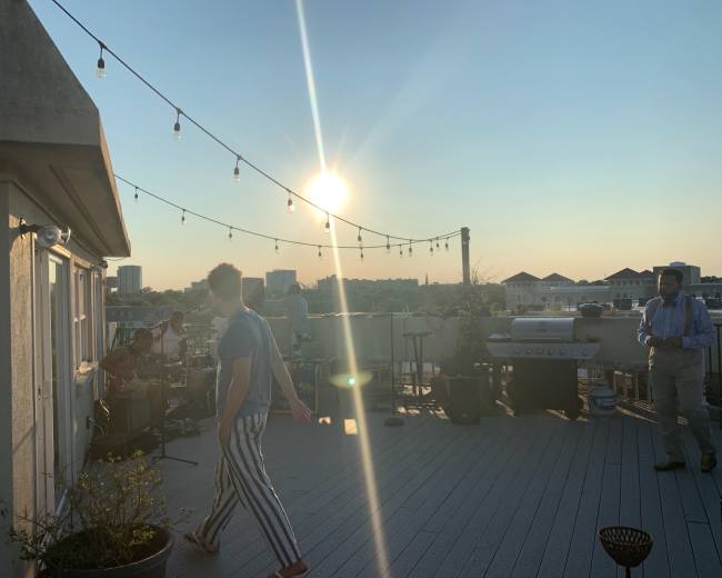 A rooftop gathering features people enjoying music under string lights with a sunset backdrop.