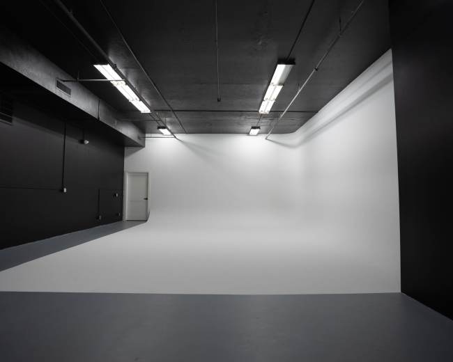 A spacious, brightly lit studio features a seamless white backdrop and a gray floor, with exposed ceiling lights illuminating the area.