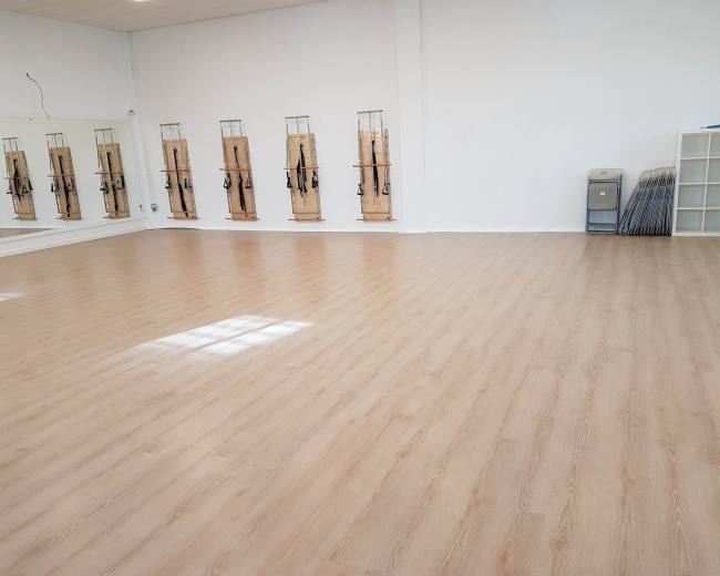 An empty fitness studio with wooden flooring, mirrors on one wall, and equipment racks along another wall.