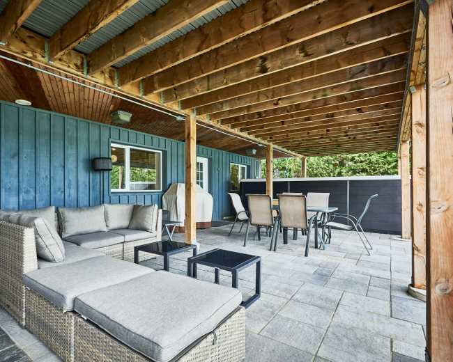 The image shows a covered patio area featuring a sectional sofa, a glass table, and dining chairs arranged on a stone surface beneath wooden beams.