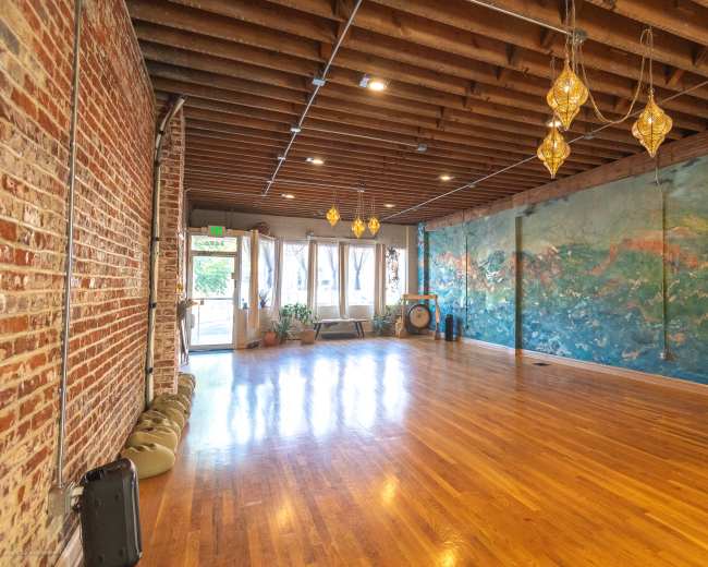 The image shows an open, spacious room with wooden flooring, exposed brick walls, and a large mural on one side, illuminated by pendant lights.
