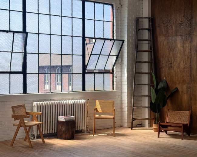 The image shows an empty, well-lit room with large windows, wooden floors, a ladder, and several simple wooden chairs arranged around a plant.