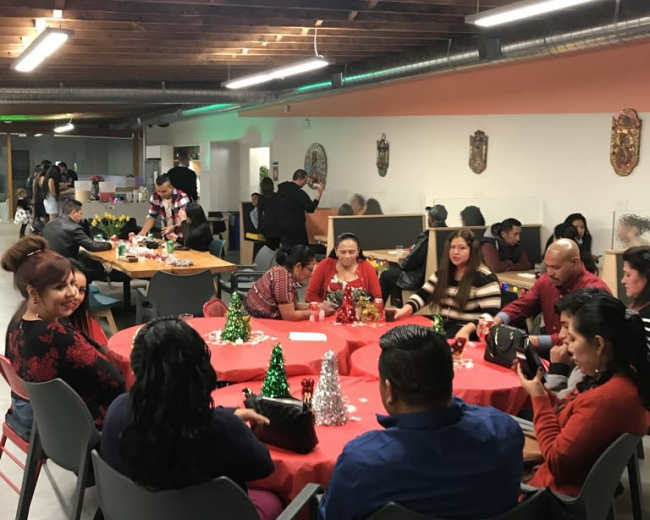 A group of people sits around decorated tables in a community gathering space, with some engaging in conversations and others arranging festive decorations.