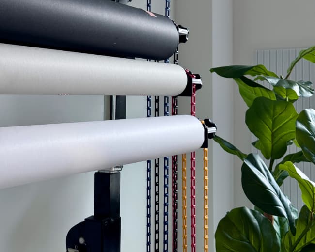 The image features a vertical display stand holding multiple rolls of colored paper alongside a potted plant in a bright room.