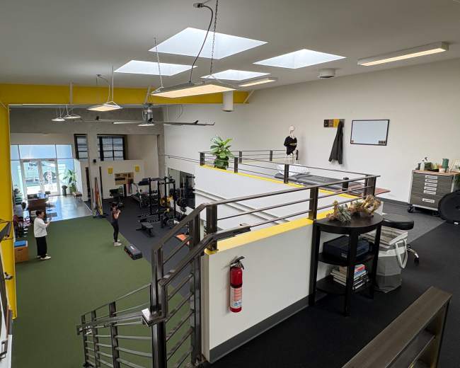 The image shows a spacious gym with various workout equipment, natural light from skylights, and a balcony area overlooking the workout space below.