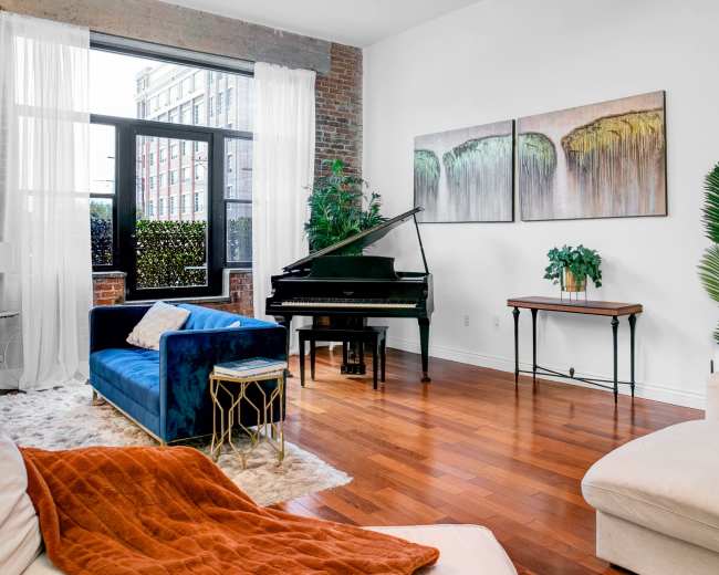 The scene features a modern living room with a grand piano, blue and beige sofas, and large windows allowing natural light to illuminate the space.