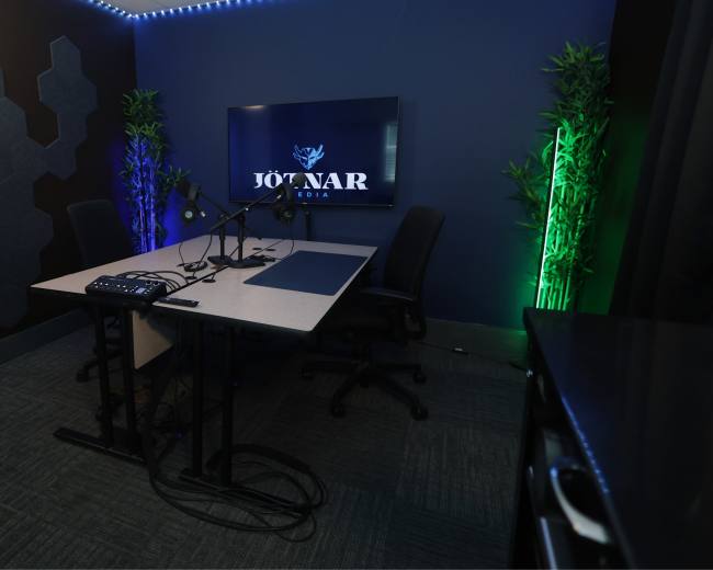 The image shows a modern podcast recording studio featuring a table with microphones, a large screen displaying the logo "JOTNAR MEDIA," and plants illuminated by colored lights.