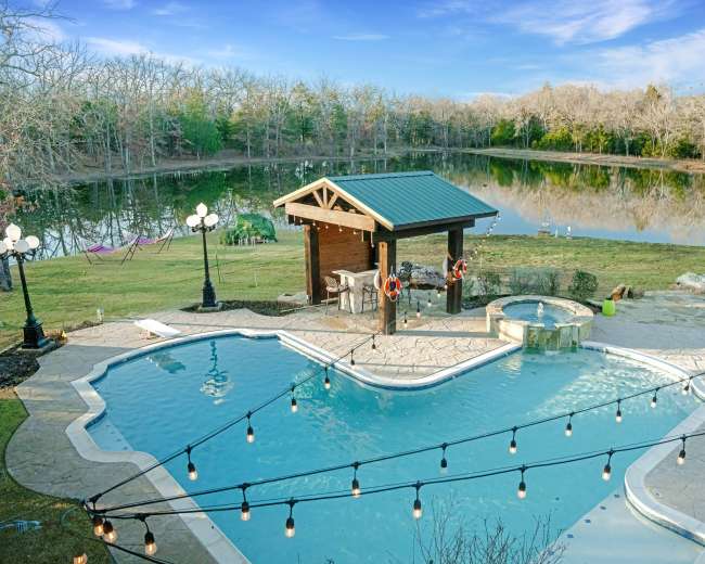 The image shows a pool area with a gazebo and a hot tub, surrounded by a landscaped yard and a lake in the background.