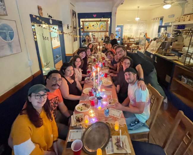 A group of people gathers around a long table in a brightly lit restaurant, sharing a meal with candles and drinks.