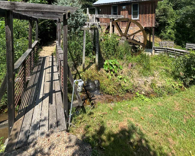 A wooden bridge leads to a rustic building with a large waterwheel, set against a backdrop of greenery and hills.