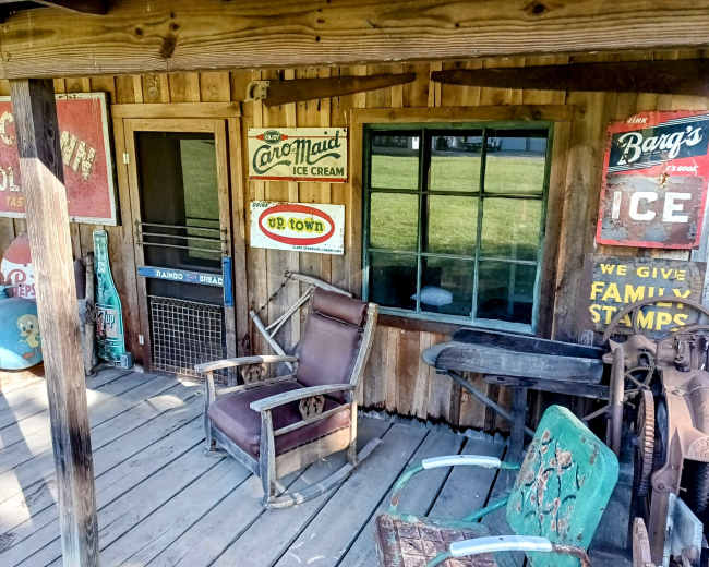 The image shows a rustic wooden building with vintage ice cream and soda advertisements, a window, and a couple of old chairs on a weathered wooden deck.
