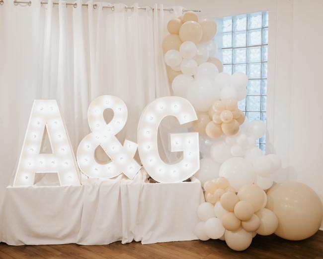 Illuminated letters "A" and "G" against a white curtain, accompanied by a decorative arrangement of balloons in neutral tones.