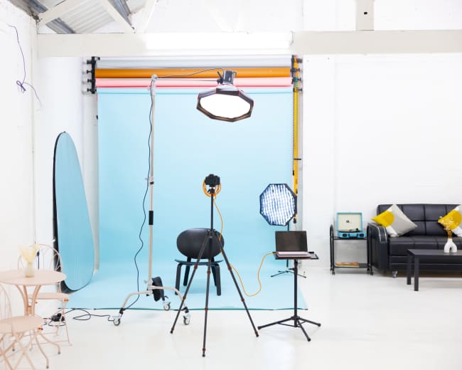 A photography studio features lighting equipment, a blue backdrop, a black chair, and a small table with chairs.