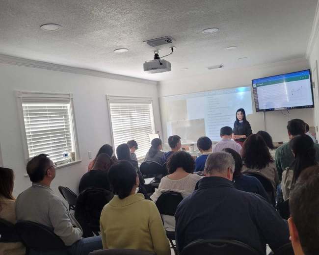 A group of people is seated in chairs, facing a woman who is presenting in front of a screen in a well-lit room.