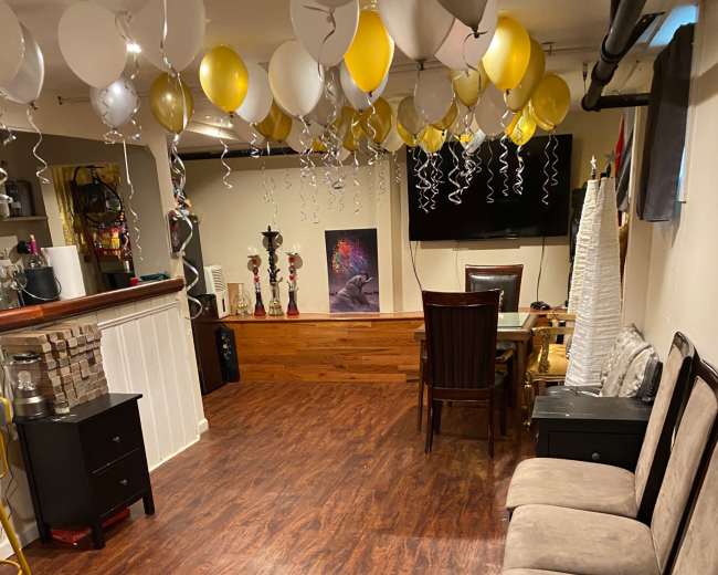 The image shows a room with wooden flooring, decorated with balloons and ribbons hanging from the ceiling, and furnished with a seating area and dining table.