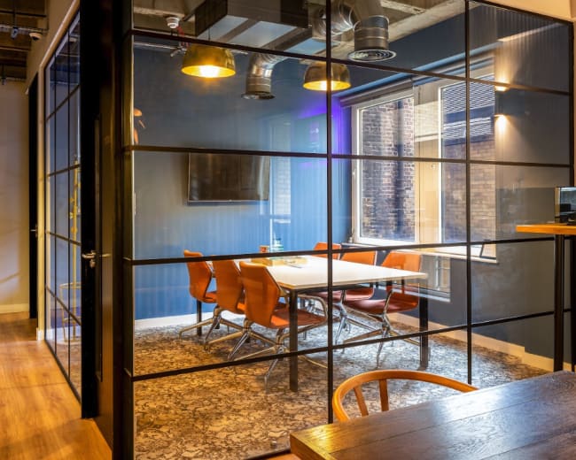 A modern conference room with a glass wall, featuring a table surrounded by leather chairs and wooden flooring.