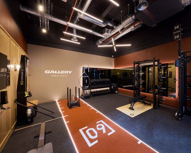 The image shows a modern fitness gym with an area for weightlifting, exercise equipment, and a mirrored wall.