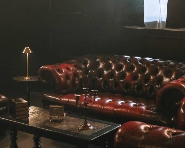 The image shows a dimly lit room featuring a red leather chesterfield sofa, an ottoman, a small side table with a lamp, and a candle holder.