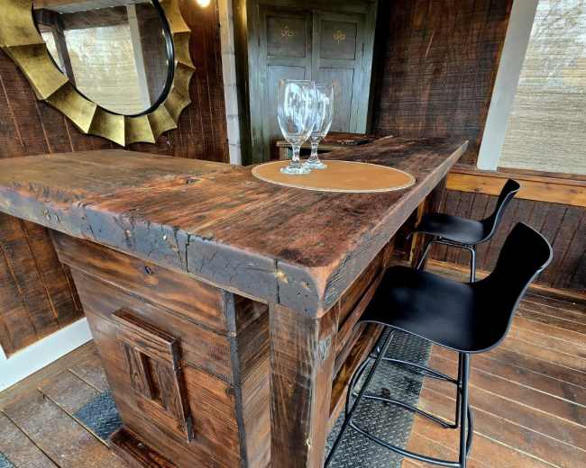 The image features a rustic wooden bar with two black stools, a mirror on the wall, and a circular placemat with two glasses on top.