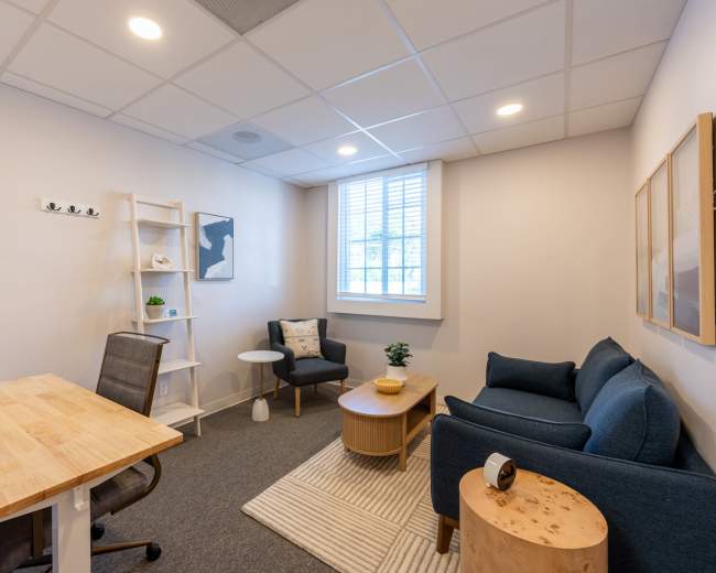 The image shows a small, modern office space featuring a desk with a chair, a coffee table, a couch, and a decorative shelf, all set against a light-colored wall.