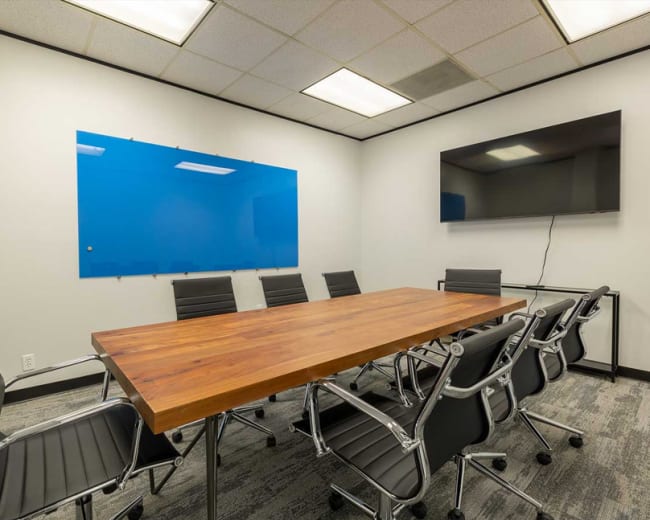 The image shows a modern conference room with a long wooden table surrounded by black office chairs, a blue whiteboard on the wall, and a wall-mounted television.