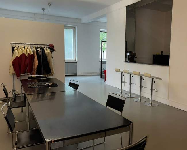 The image shows a spacious room with a long table, several bar stools at a counter, a clothing rack with coats, and a large window providing natural light.