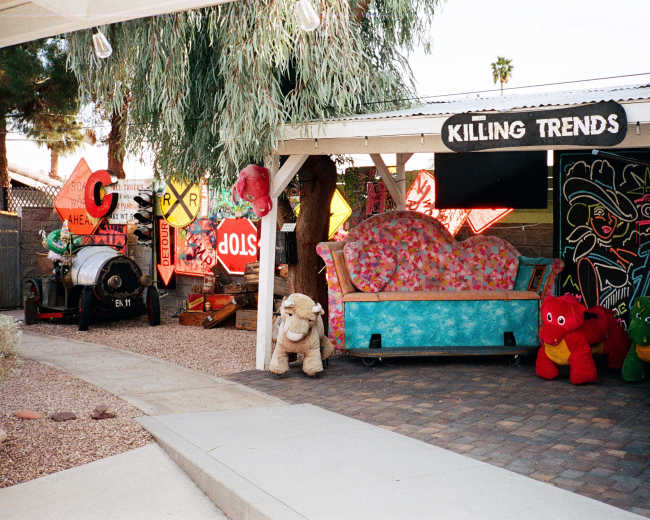 The image depicts a colorful outdoor space featuring a blue patterned couch, stuffed animals, quirky signage, and an antique car amidst gravel and potted plants.