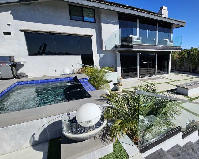 The image shows a modern outdoor space featuring a pool with a spa, landscaped areas, and a two-story house with large glass windows.